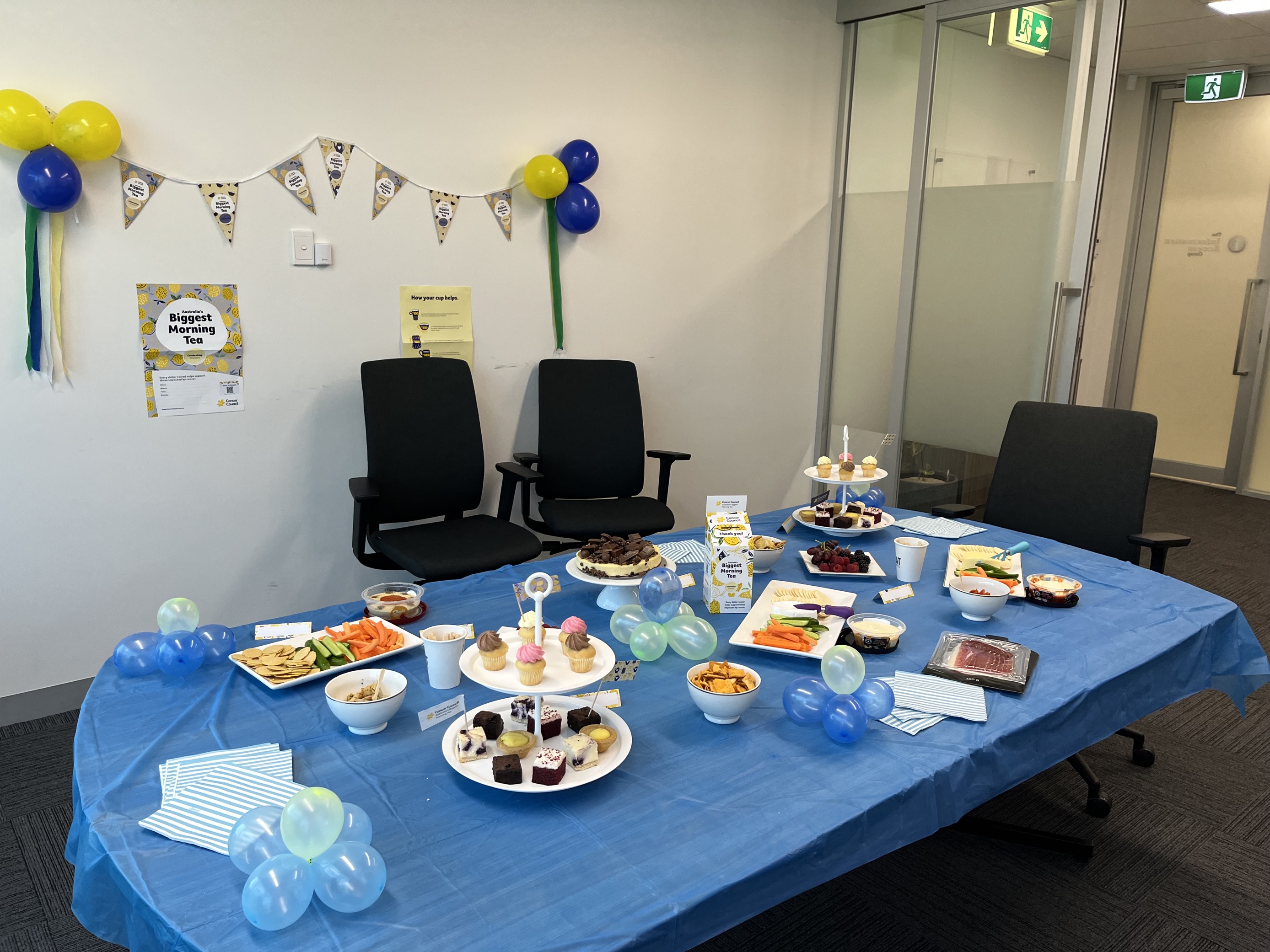 The Information Access Group conference room table decorated with blue and yellow balloons and a blue table cloth. On the table are a variety of food and snacks, including cupcakes, dips, fruits, and biscuits. On the wall behind is a poster that says 'Australia's Biggest Morning Tea'. Click to view full image.