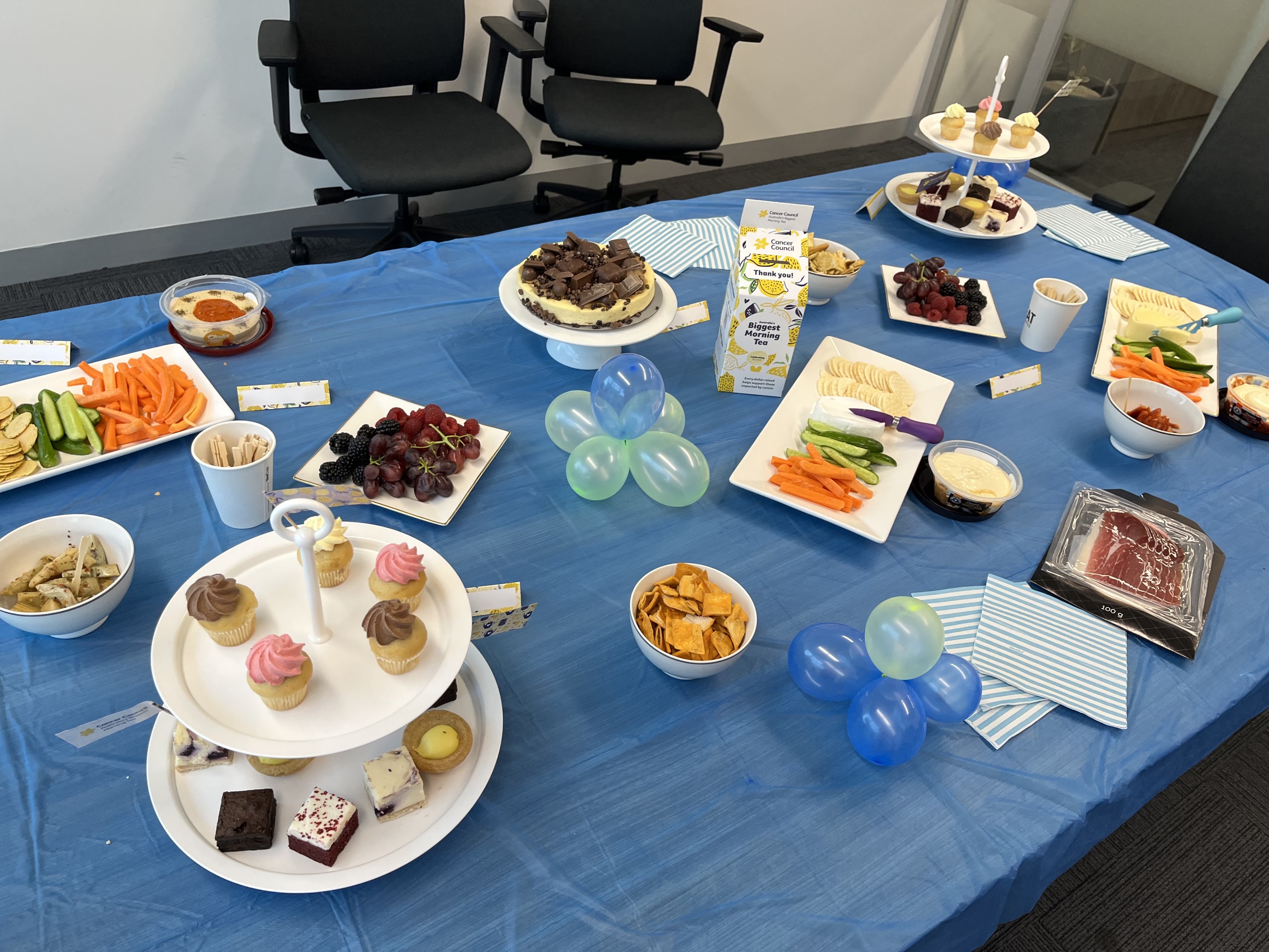 A closer shot of the conference room table decorated with blue and yellow balloons and a blue table cloth. On the table are a variety of food and snacks, including cupcakes, dips, fruits, and biscuits. Click to view full image.