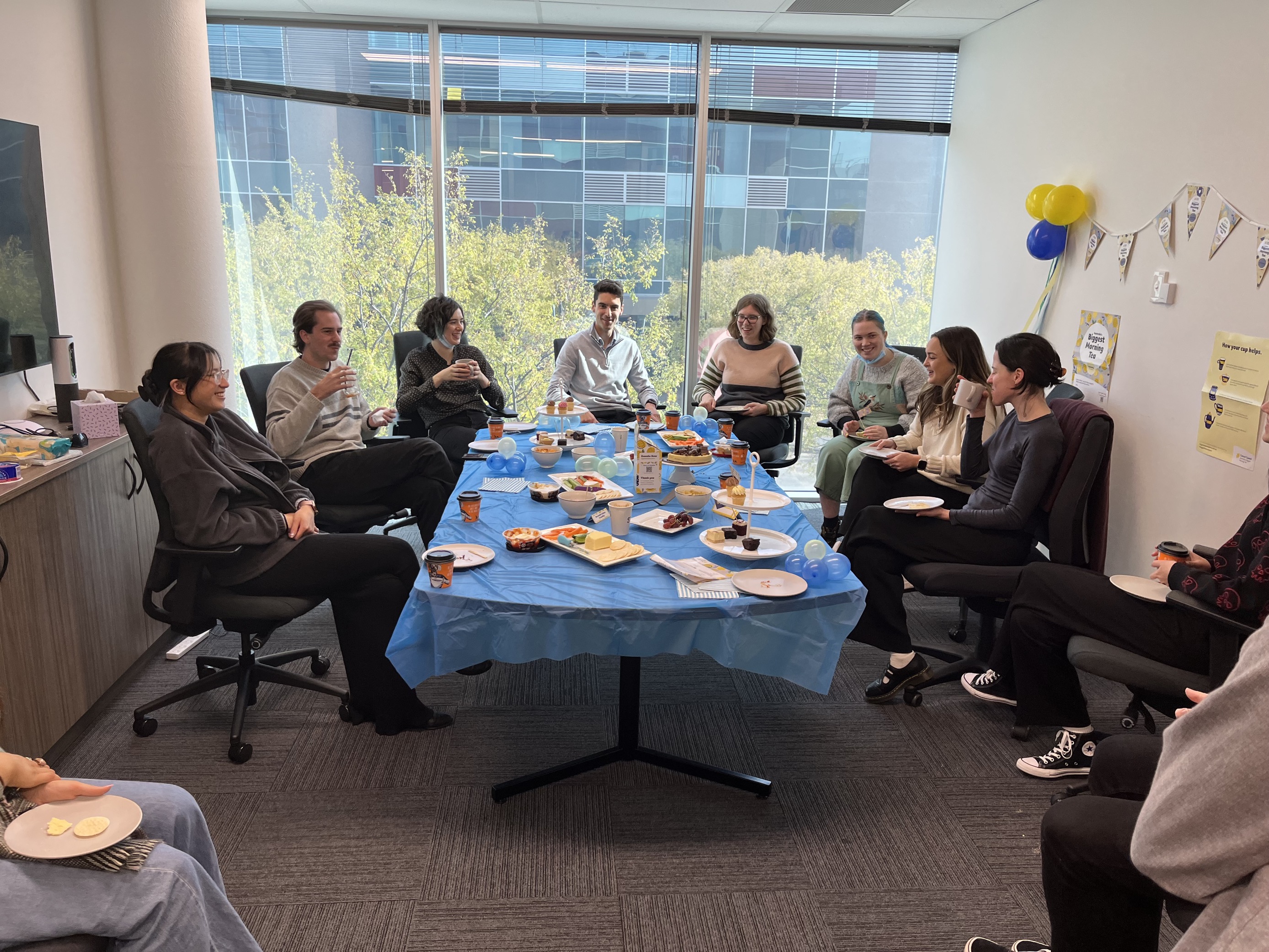 People from the Information Access Group sitting around the conference table, celebrating Australia's Biggest Morning Tea, eating food and drinking tea and/or coffee. Some are holding mugs, and some are holding plates. Pictured in the photograph are Fiona, Tom L, Annie, Liam, Renee, Zoe, Taylah, and Evelyn. Click to view full image.