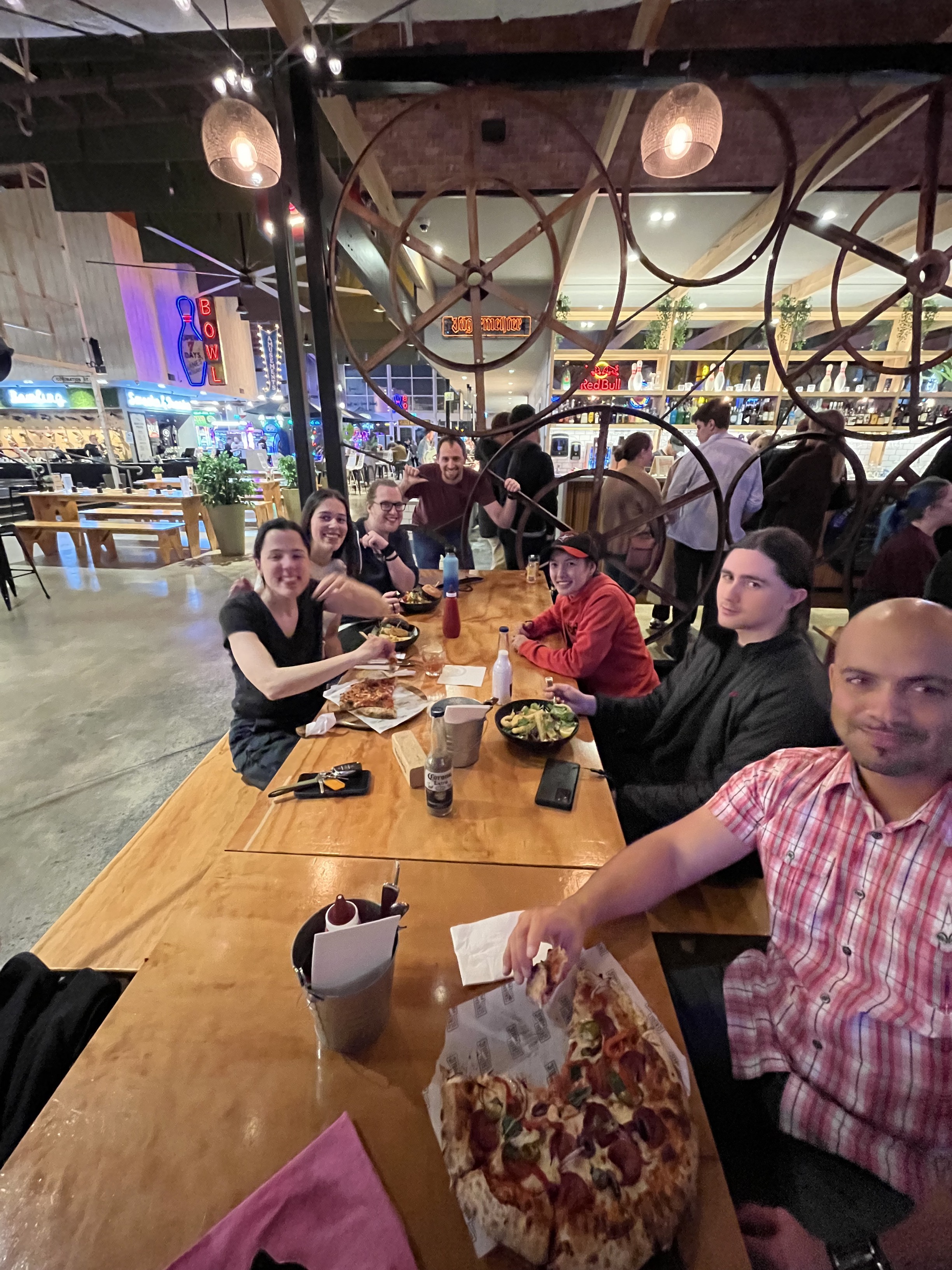 People from the Information Access Group sitting at a table together at the bowling venue. On the table are pizzas, pasta, and drinks. Pictured in the photograph are Evelyn, Sienna, Tom M, David M, Shannon, Jason, and Sean. They are smiling at the camera. Click to view full image.