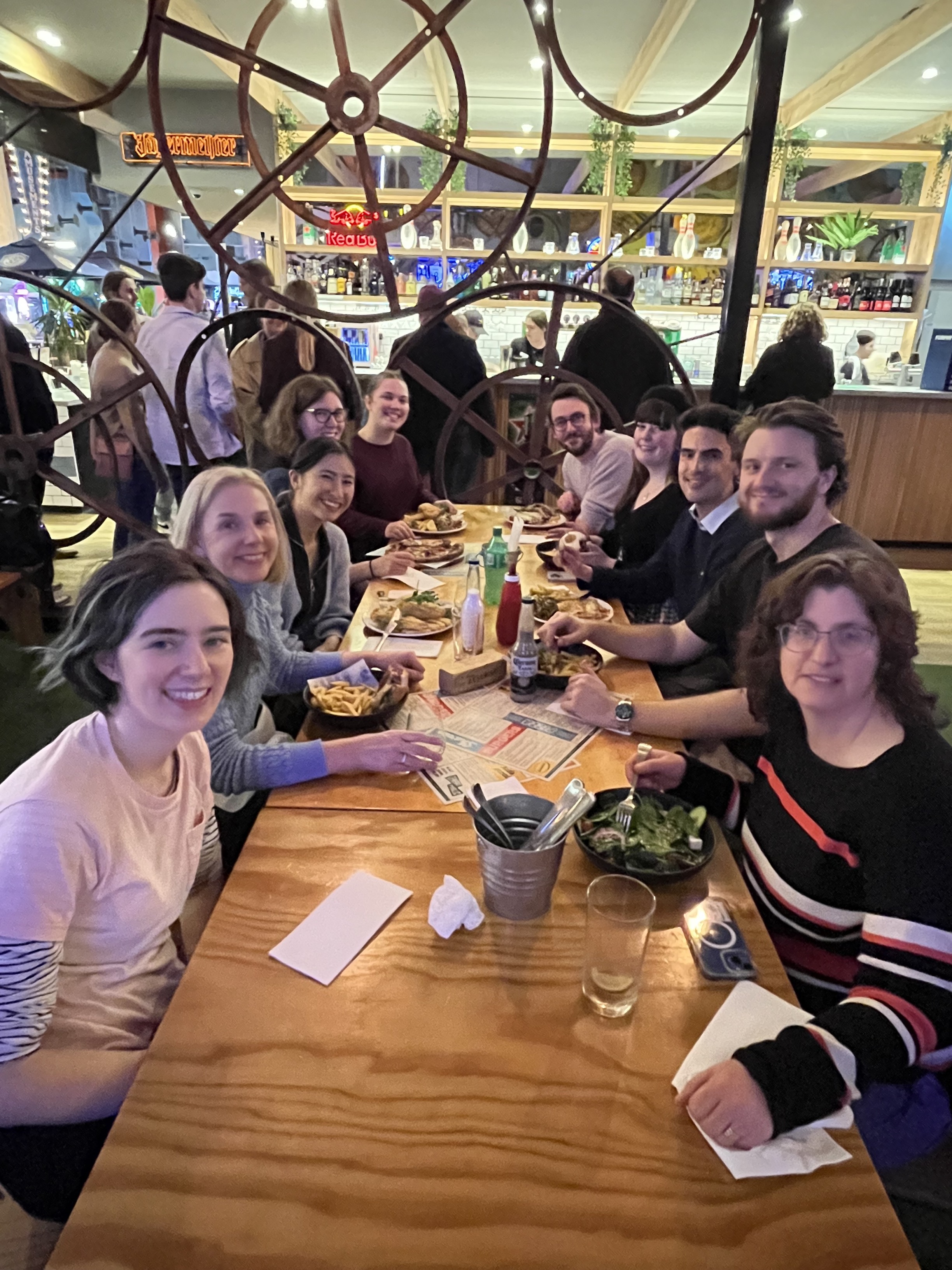 People from the Information Access Group sitting at a table together at the bowling venue. On the table are salads, chips, and burgers. Pictured are Annie, Sally, Fiona, Renee, Zoe, Larry, Mikayla, Liam, Fraser, and Hilary. They are smiling at the camera. Click to view full image.