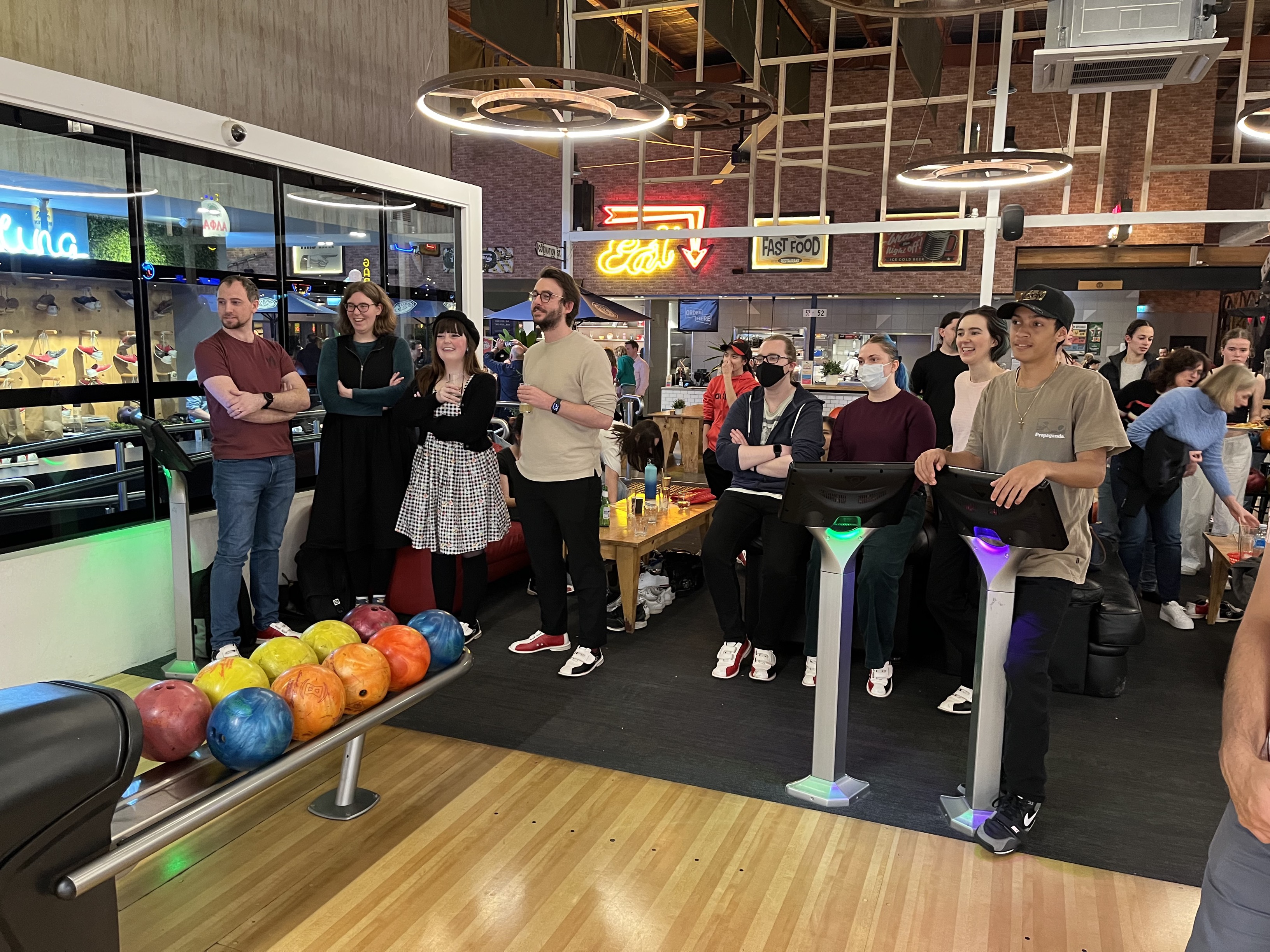 People from the Information Access Group at a bowling alley watching someone bowl off camera. In front of them are bowling balls and monitors for scoring. Pictured in the foreground are David M, Renee, Mikayla, Larry, Tom M, Zoe, Annie, and Malachi. Click to view full image.