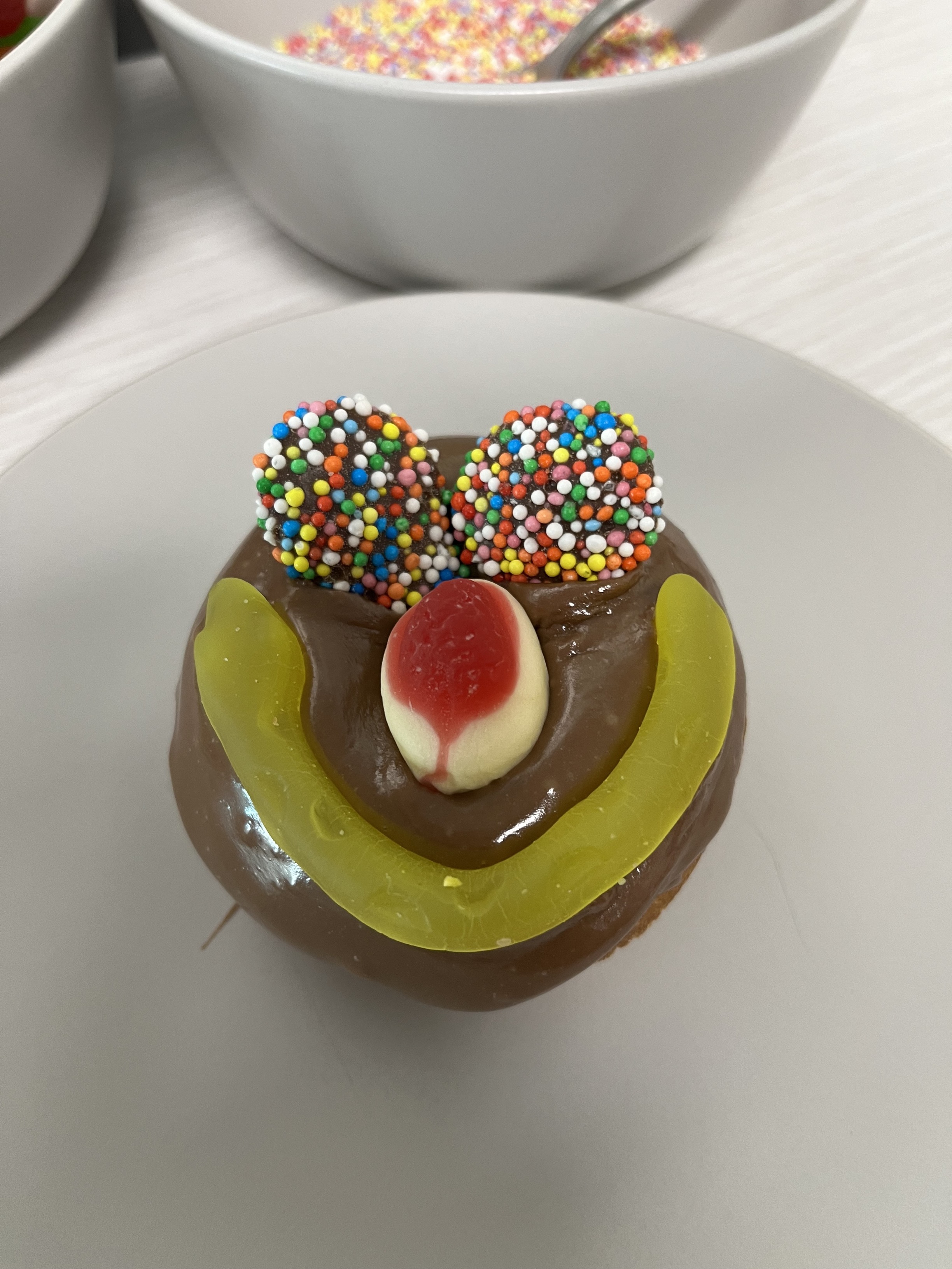 A donut decorated as a smiley face. Chocolate icing is used as the base, with two chocolate freckles for the eyes, a strawberry and cream lolly for the nose, and a yellow snake as the smile. Click to view full image.