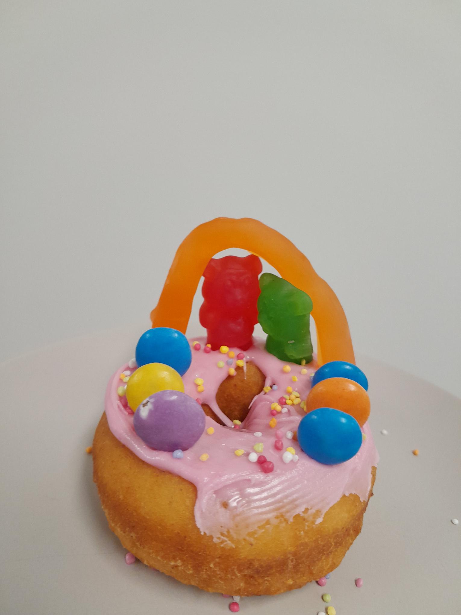 A donut decorated as a scene of a wedding. Red and green gummy bears are getting married in front of an orange snake used as an arch. Smarties are the witnesses. The base is strawberry icing with sprinkles. Click to view full image.