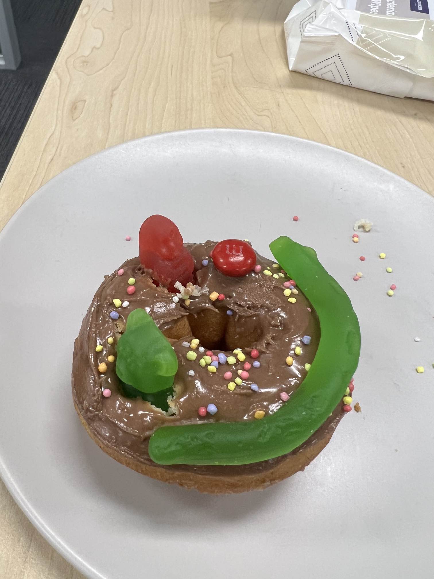 A donut decorated as a smiley face. The base is chocolate icing with sprinkles, with red and green gummy bears for the eyes and a green snake as the smile. Click to view full image.