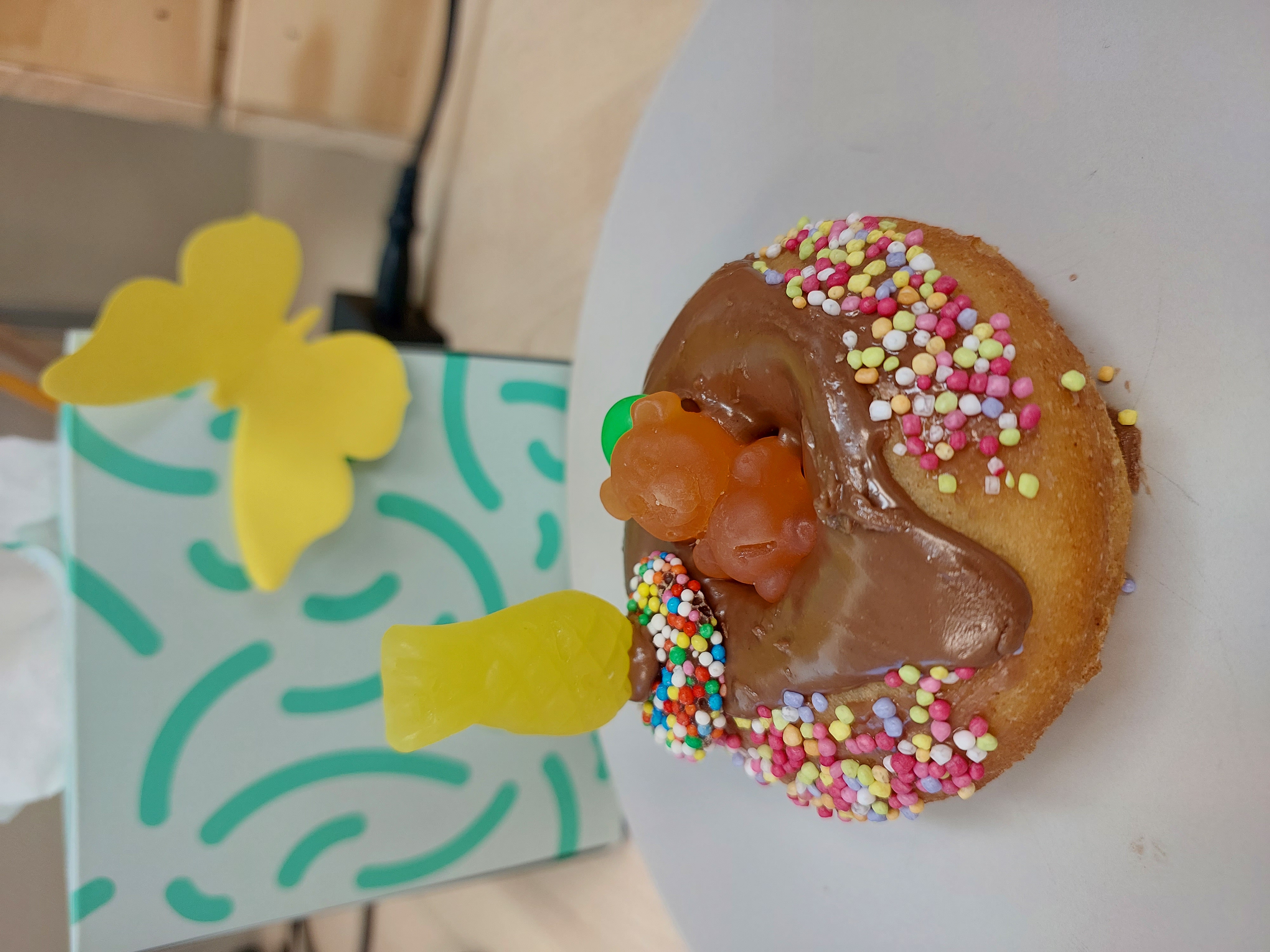 A donut decorated as a scene of an orange gummy bear relaxing in a donut ring in a pool. The base is chocolate icing and sprinkles. A gummy pineapple is stood on a freckle next to gummy bear on the donut. Click to view full image.