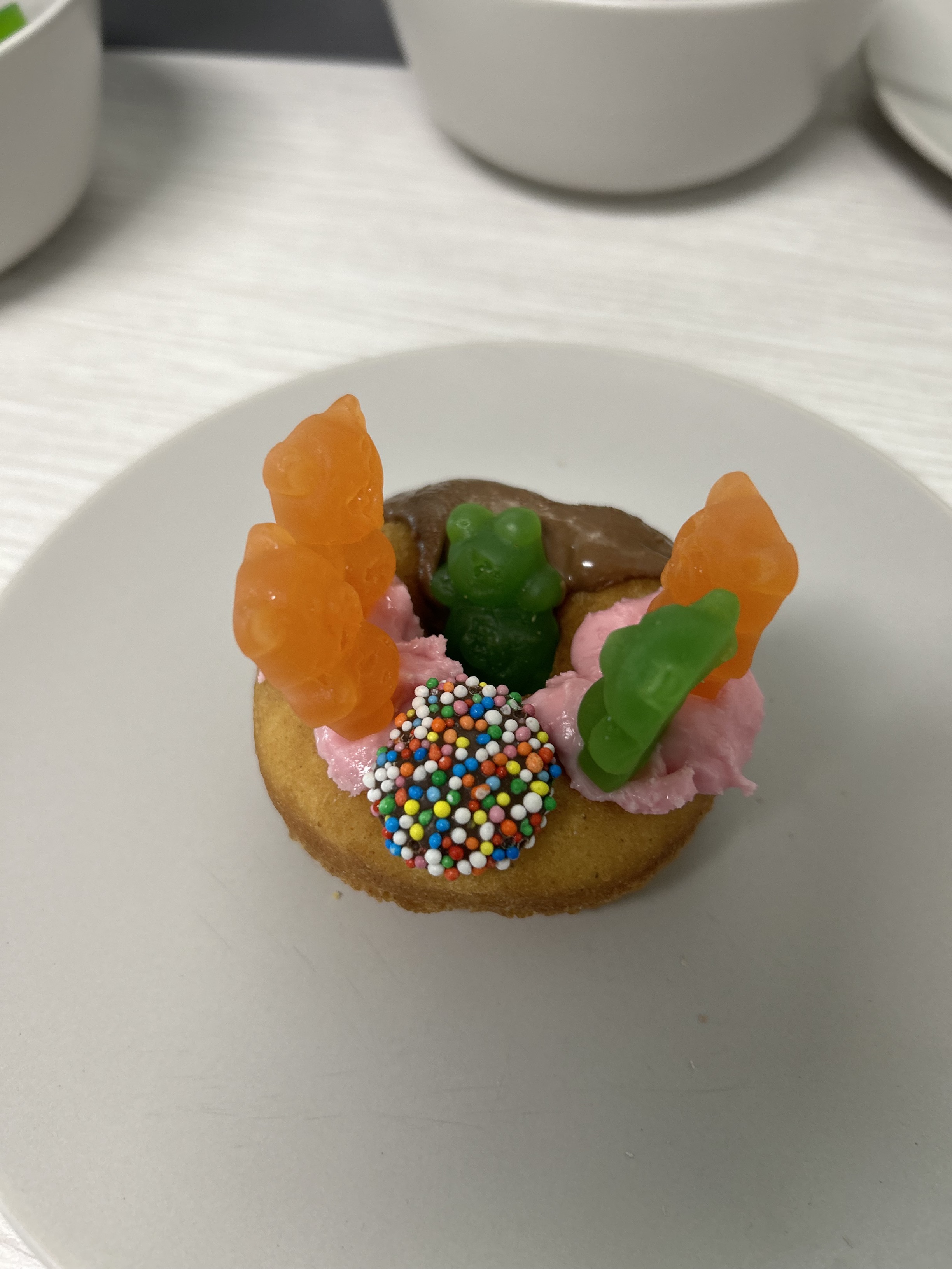 A donut decorated with chocolate and strawberry icing. Four gummy bears are stood on top of the donut, and one gummy bear is in the donut hole. A freckle is also used for decoration. Click to view full image.