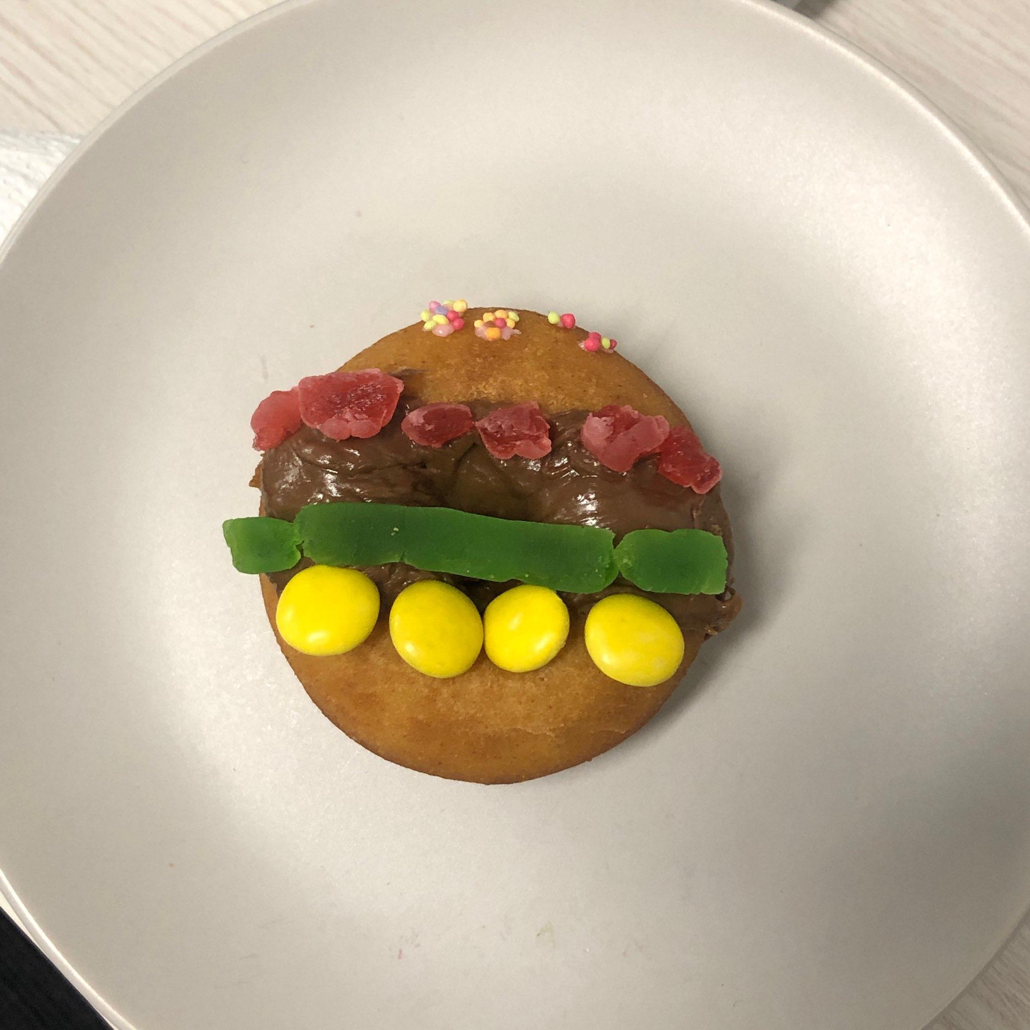 A donut decorated to look like a burger using a red gummy bear that is chopped up, a green snake, chocolate icing, yellow smarties, and sprinkles. Click to view full image.