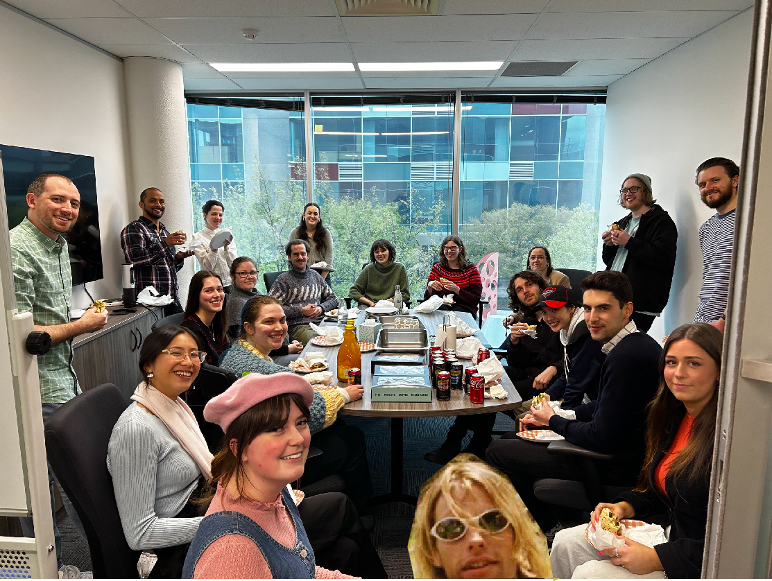 People from the Information Access Group in the conference room smiling at the camera, celebrating Eat Outside Day. Some people are holding their sandwich they are having for lunch. On the table are donuts and soft drinks. Pictured in the photograph are Mikayla, Fiona, David M, Sean, Evelyn, Zoe, Sienna, Laura, Tom L, Taylah, Annie, Renee, Kellie, Tom M, Fraser, Edward, Shannon, Liam, and Savanah. Stephen is photoshopped into the foreground with blonde hair and sunglasses. Click to view full image.