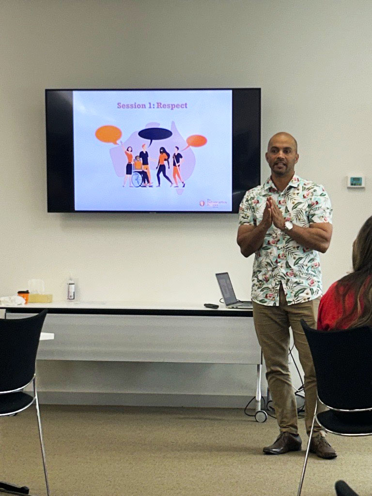 Sean presenting at the Information Access Group values session. Behind him is a PowerPoint slide on a TV. The text reads 'Session 1: Respect'. Click to view full image.