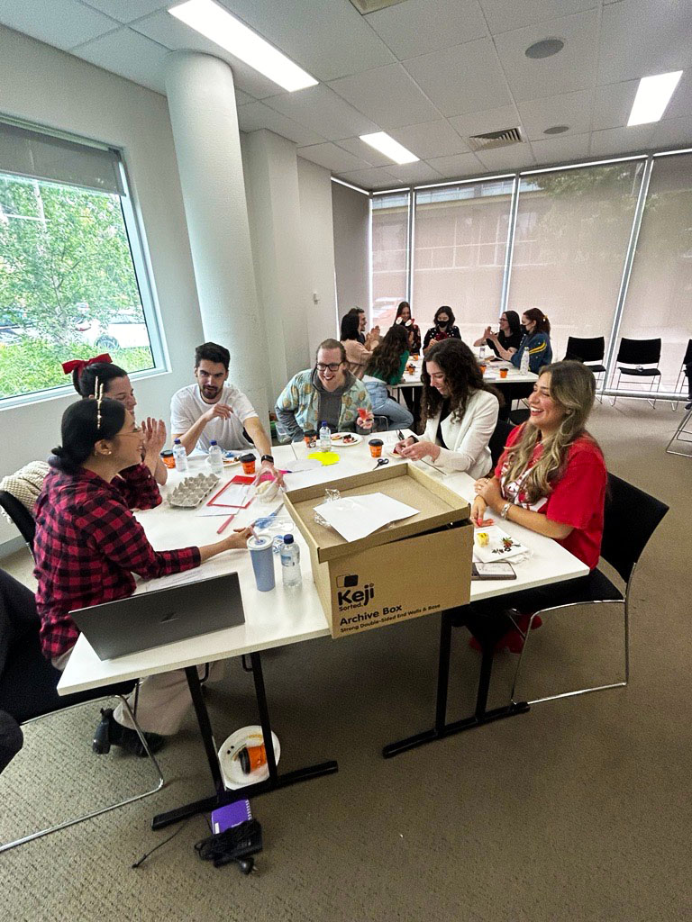 People from the Information Access Group sitting in groups at multiple tables in a conference room, taking part in the Information Access Group values session. On the tables are plates of food and different craft supplies. People are using the craft supplies to create dioramas. Pictured in the photograph are Fiona, Evelyn, Liam, Tom M, Catie, and Savanah. Click to view full image.