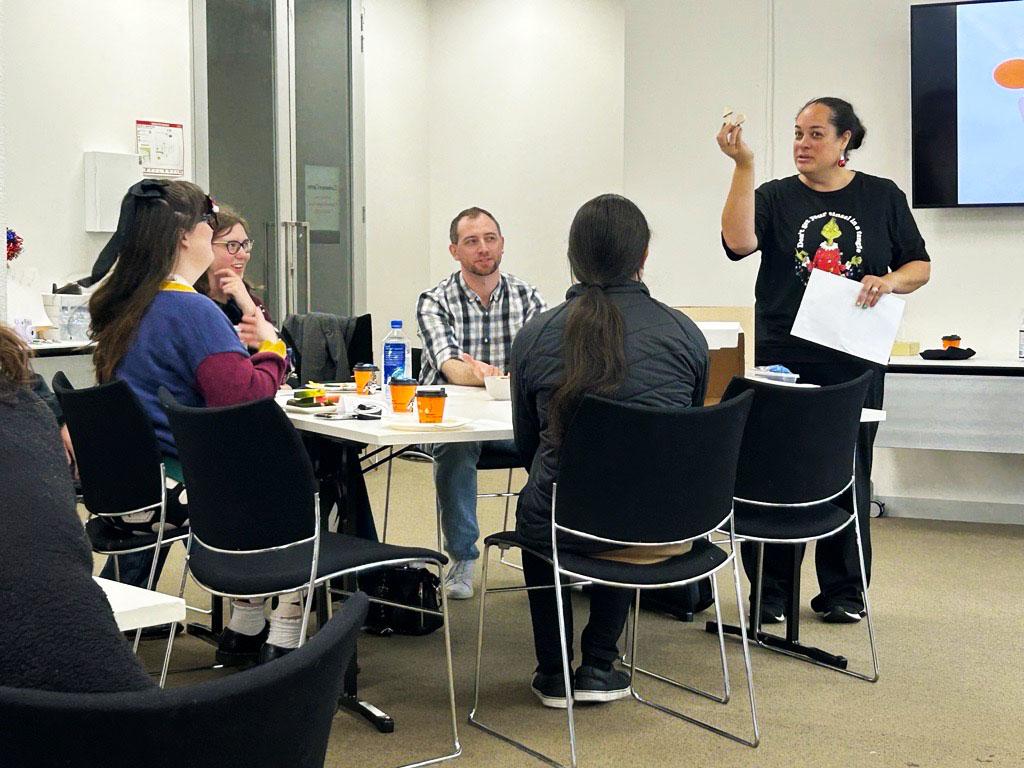 Lynn holding up craft supplies to explain an activity at the Information Access Group values session. She is standing next to a table, where people are sitting and listening to her. Other people pictured in the photograph are Mikayla, Renee, David M, and Jason. Click to view full image.