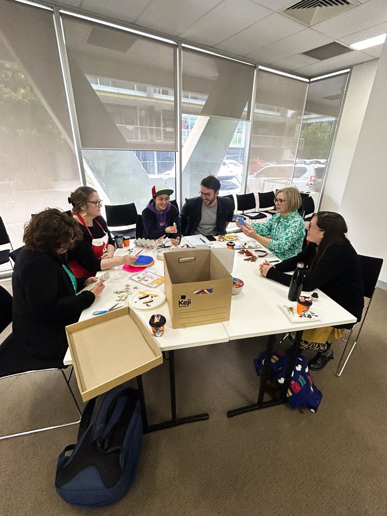 People from the Information Access Group sitting at a table in a conference room, taking part in the Information Access Group values session. On the table are plates of food and different craft supplies. People are using the craft supplies to create a diorama. Pictured in the photograph are Hilary, Laura, Shannon, Larry, Sally, and Melanie. Click to view full image.